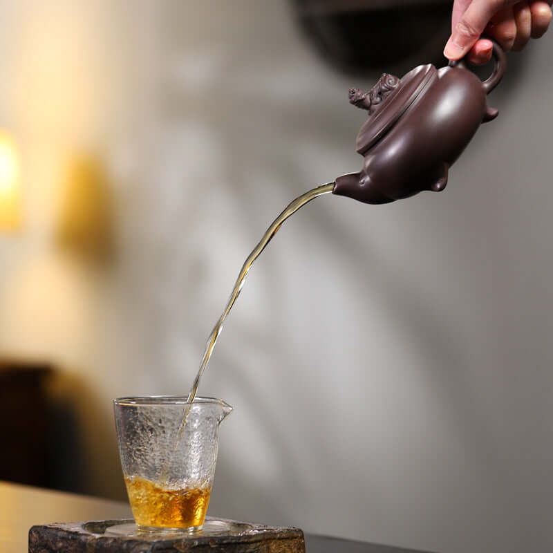 Handmade Yixing purple sand teapot pouring tea into a glass cup, showcasing elegant craftsmanship and traditional tea art.