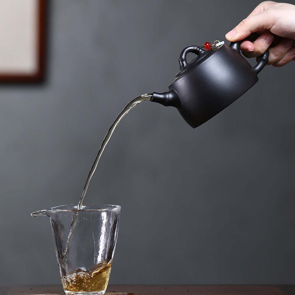 Handmade Yixing purple sand teapot pouring tea into a glass cup, showcasing original craftsmanship and elegance.