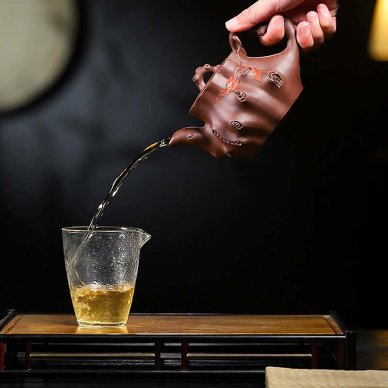 Handmade Yixing purple sand teapot pouring tea into a glass cup on a wooden tray, showcasing fine craftsmanship.