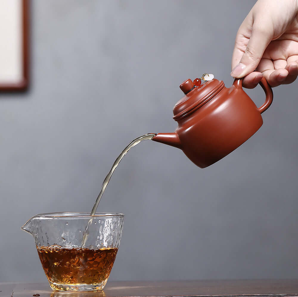 Handmade Yixing purple sand teapot pouring tea into glass cup, showcasing traditional Kung Fu tea ceremony.
