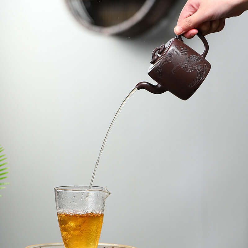 Handmade Yixing purple sand teapot pouring tea into a glass cup, showcasing its elegant design and craftsmanship.