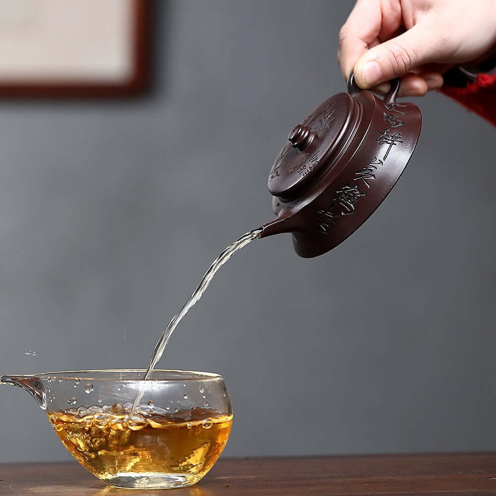 Handmade Yixing purple sand teapot pouring tea into a glass cup, showcasing traditional craftsmanship and elegance.