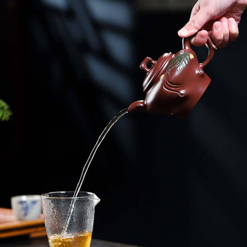Handmade Yixing purple sand teapot pouring tea into a glass cup, showcasing elegant craftsmanship.