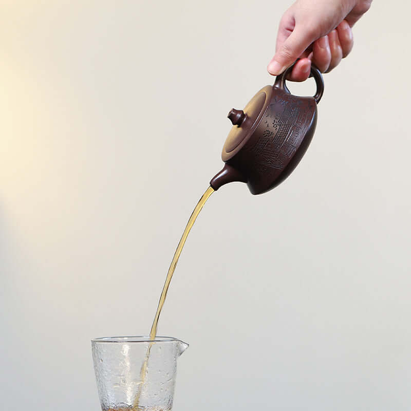 Hand pouring tea from a handmade Yixing purple sand teapot into a clear cup, showcasing elegant design and craftsmanship.