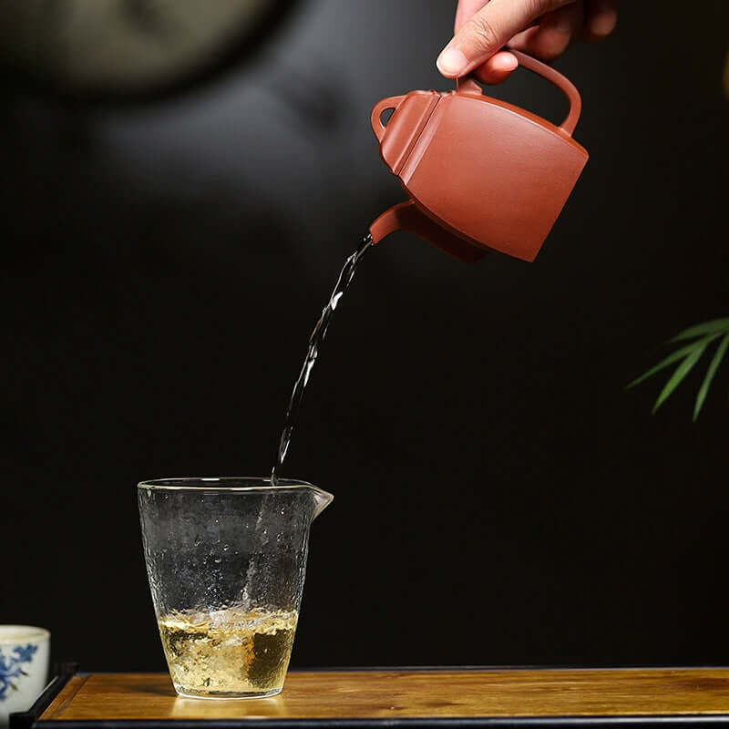 Handmade Yixing purple sand teapot pouring tea into a glass cup, showcasing traditional tea craftsmanship.
