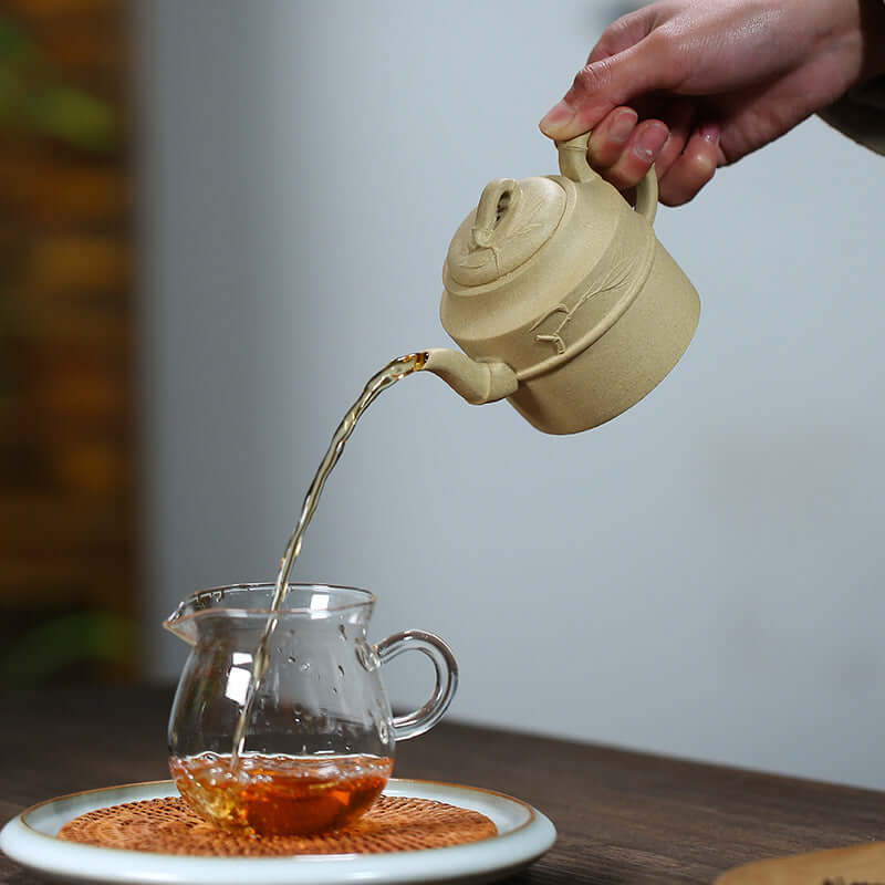 Handmade Yixing purple sand teapot pouring tea into a glass cup on a wooden table, showcasing traditional Kung Fu tea set.