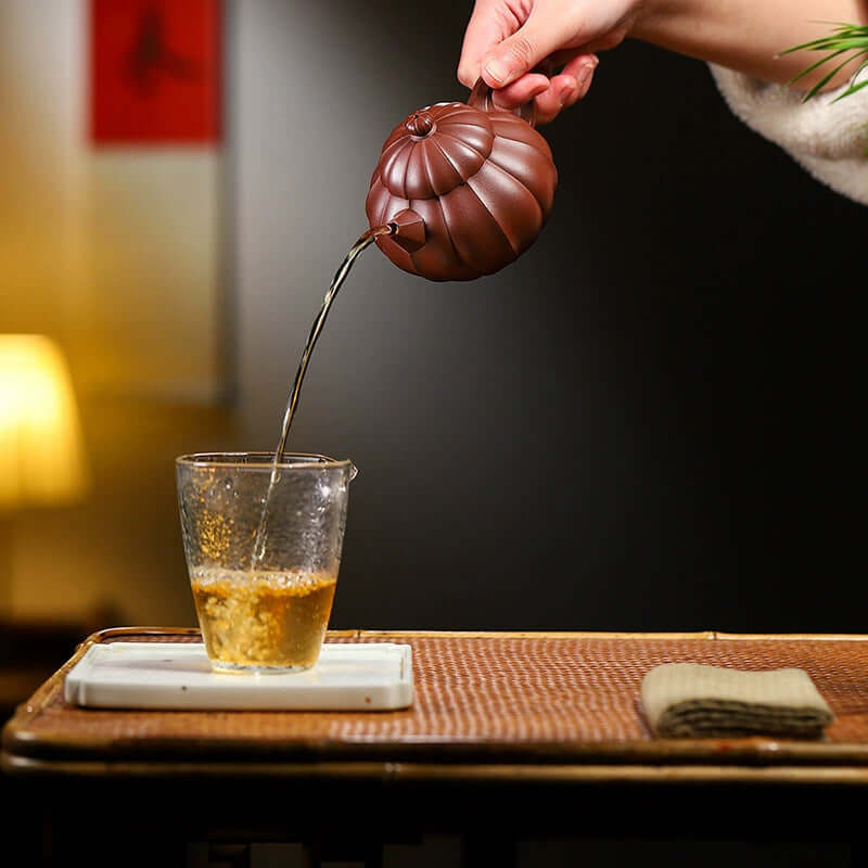 Handmade Yixing purple sand teapot pouring tea into a glass cup on a wooden tray, showcasing fine craftsmanship.