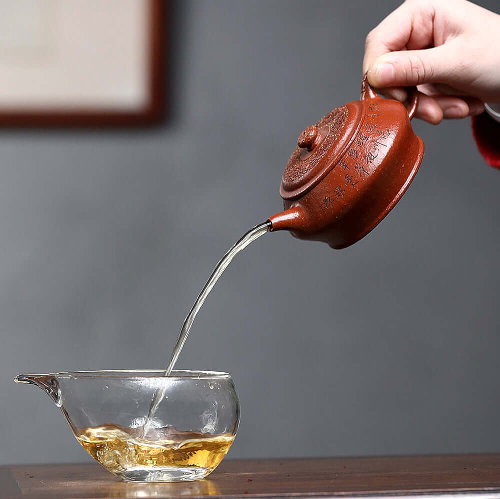 Handmade Yixing purple sand teapot pouring tea into a glass cup, showcasing craftsmanship and elegance.