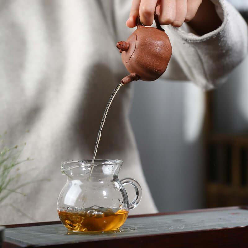 Handmade Yixing purple sand teapot pouring tea into a glass cup, showcasing artisanal craftsmanship.