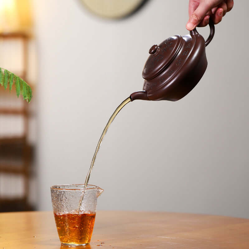 Handmade Yixing purple sand teapot pouring tea into a glass cup, showcasing craftsmanship and elegance.