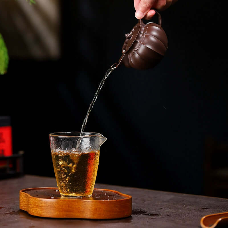 Handmade Yixing purple sand teapot pouring tea into a glass cup on a wooden tray, showcasing original craftsmanship.