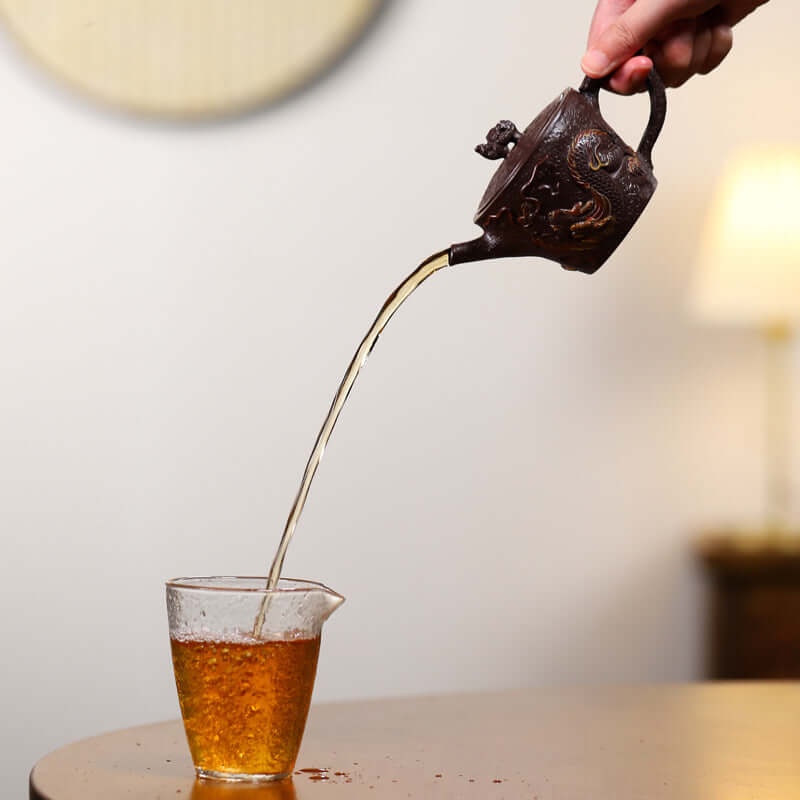 Hand pouring tea from a handmade Yixing purple sand teapot into a glass cup, showcasing tea's rich color.