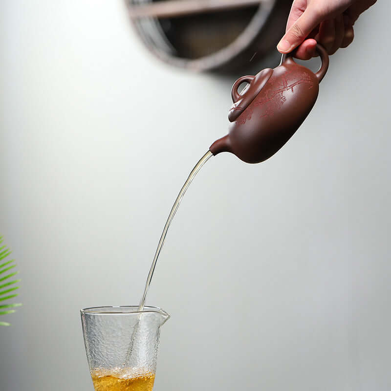 Handmade Yixing purple sand teapot pouring tea into a glass cup, showcasing elegant craftsmanship and functionality.