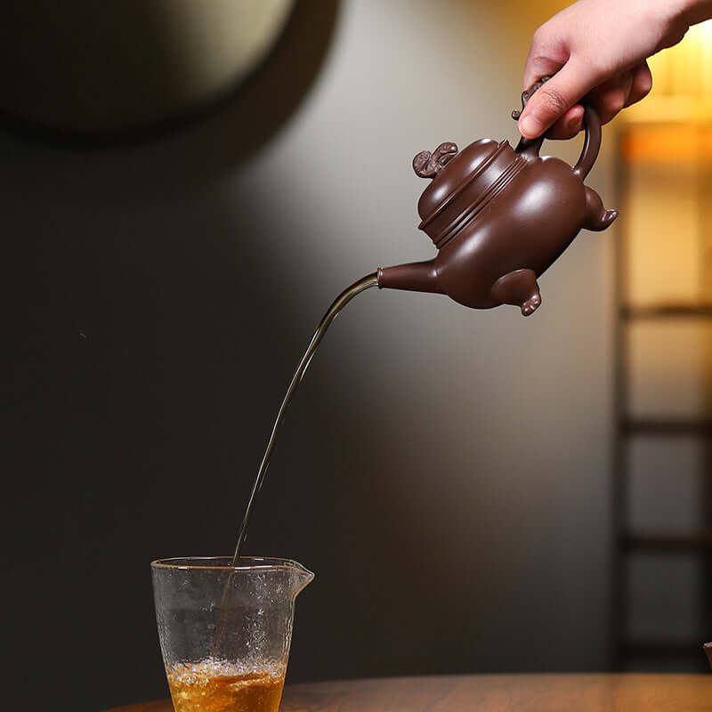 Handmade Yixing purple sand teapot pouring tea into a glass cup, showcasing elegant design and craftsmanship in low light.