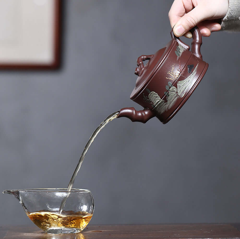 Handmade Yixing purple sand teapot pouring tea into glass cup, showcasing unique painted landscape and bamboo design.