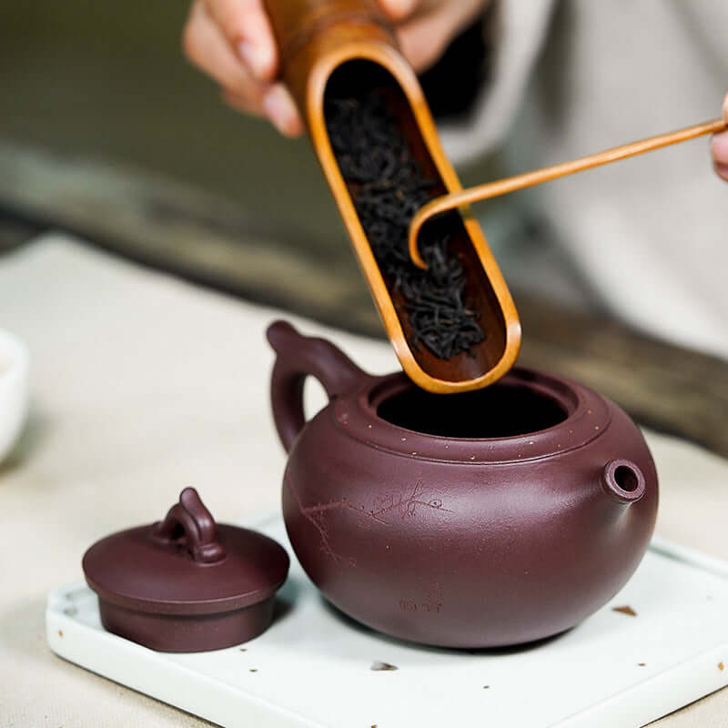 Handmade Yixing purple sand teapot with loose tea being added, showcasing traditional tea preparation.