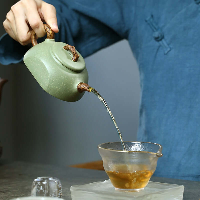 Handmade Yixing purple sand teapot pouring tea into a glass cup, showcasing traditional craftsmanship and elegance.
