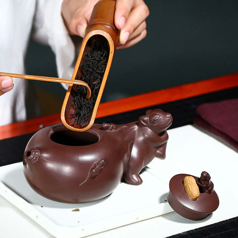 Handmade Yixing purple sand teapot with intricate mouse design, filled with loose black tea leaves.