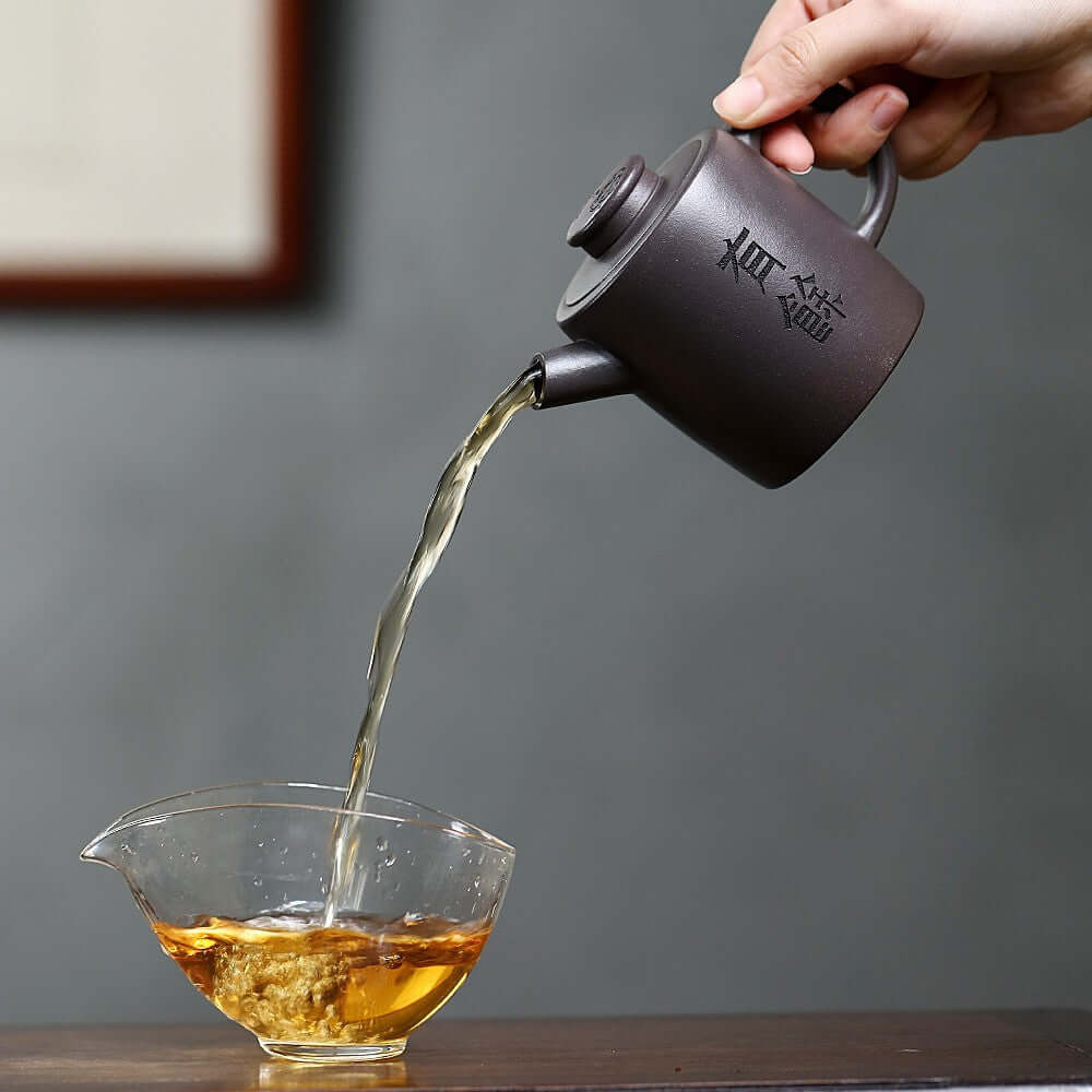 Handmade Yixing purple sand teapot pouring tea into a glass cup, showcasing artisan craftsmanship.
