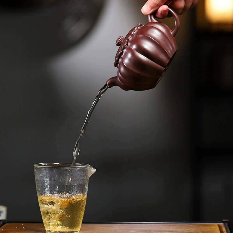 Handmade Yixing purple sand teapot pouring tea into a glass cup, showcasing craftsmanship and original purple sand.