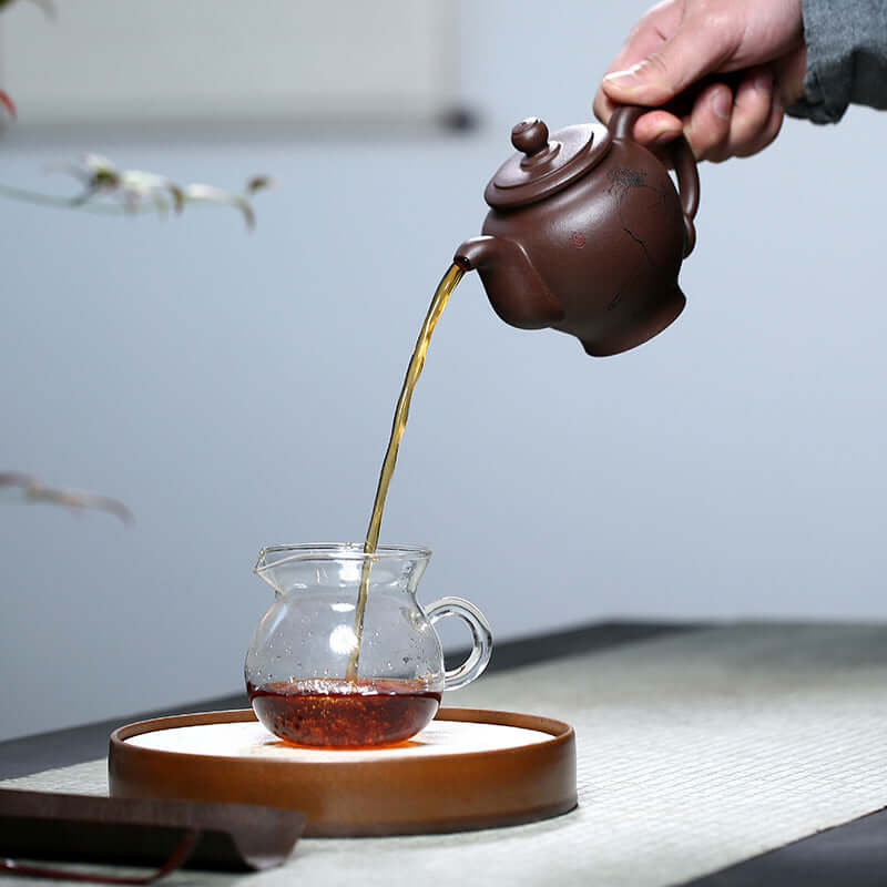 Handmade Yixing purple sand teapot pouring tea into a glass cup on a traditional tea set.
