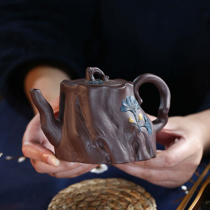 Handmade Yixing purple sand teapot shaped like a ginkgo tree stump, featuring intricate details, held by a person.