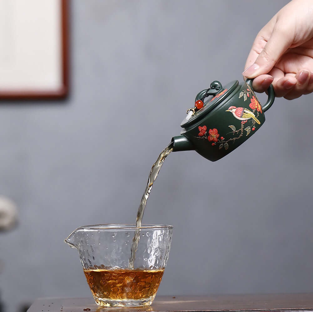 Handmade Yixing purple sand teapot pouring tea into a glass cup, showcasing intricate painted design and craftsmanship.
