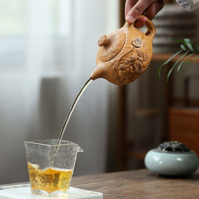 Handmade Yixing purple sand teapot pouring tea into a glass cup, showcasing craftsmanship and elegance.