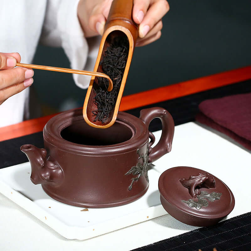 Handmade Yixing purple sand teapot being filled with loose tea leaves using bamboo scoop.