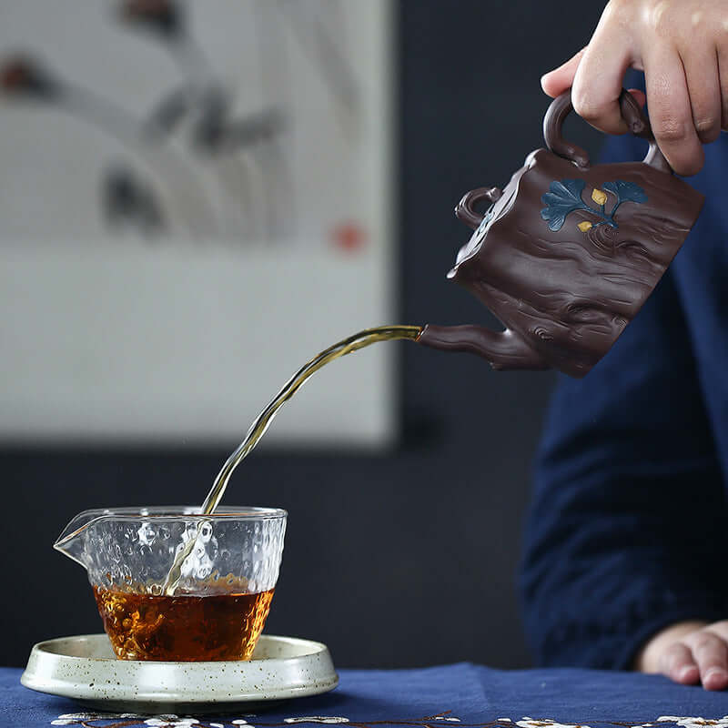 Handmade Yixing purple sand teapot pouring tea into a transparent cup on a table.