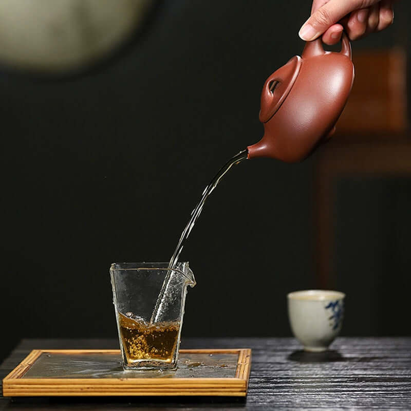 Handmade Yixing purple sand teapot pouring tea into a glass cup on a bamboo tray.