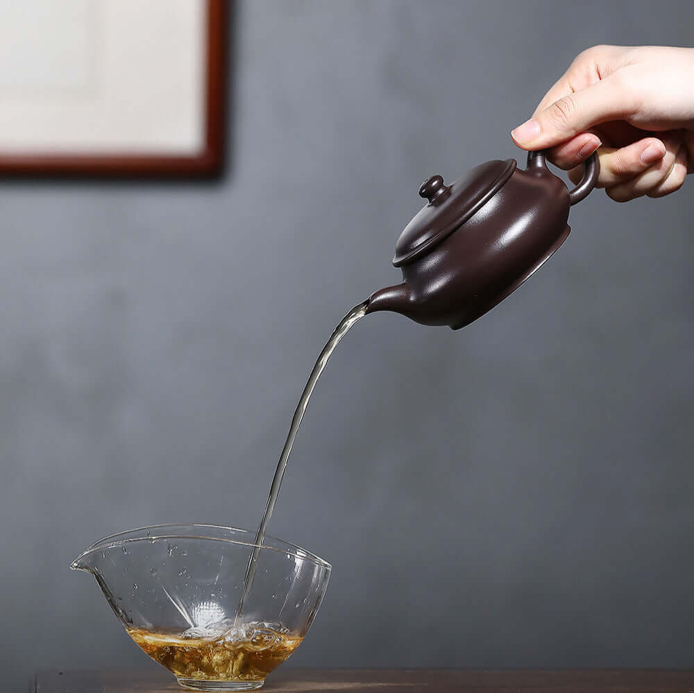 Handmade Yixing purple sand teapot pouring tea into a glass cup, showcasing craftsmanship and elegance for Kung Fu tea.