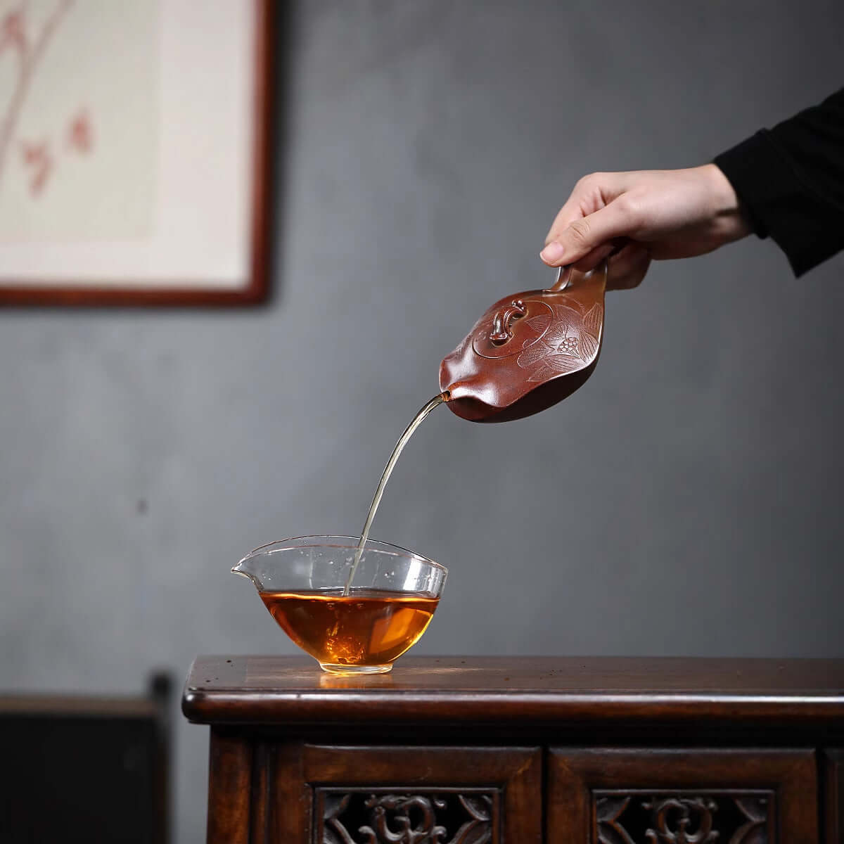 Hand pouring tea from a handmade Yixing purple sand teapot into a glass cup on a wooden table.