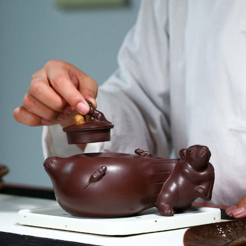 Handmade Yixing purple sand teapot with cute mouse design, showing a craftsman elegantly placing the lid.