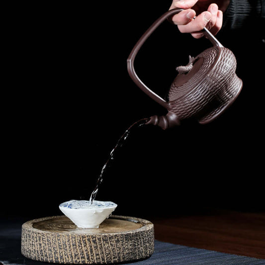 Handmade Yixing purple sand teapot pouring water into an elegant teacup on a wooden tray.