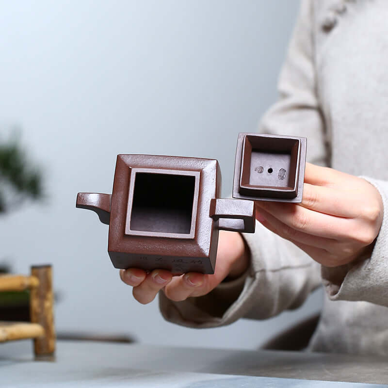 Handmade Yixing purple sand teapot held by a person, showcasing its unique square design and inner compartment.