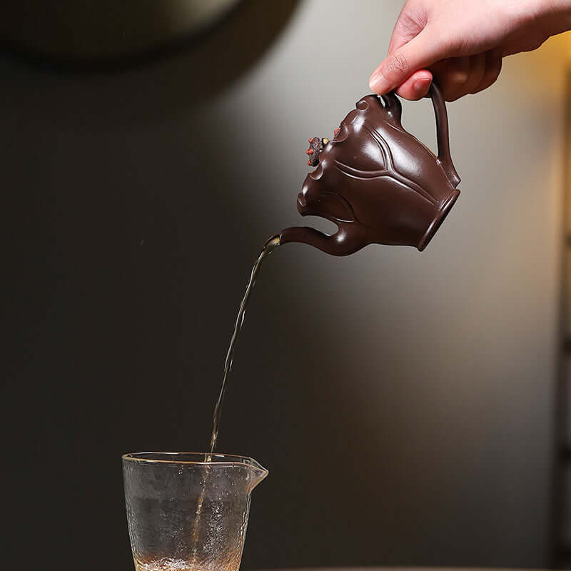 Handmade Yixing purple sand teapot pouring tea into a glass cup, showcasing unique craftsmanship and design.