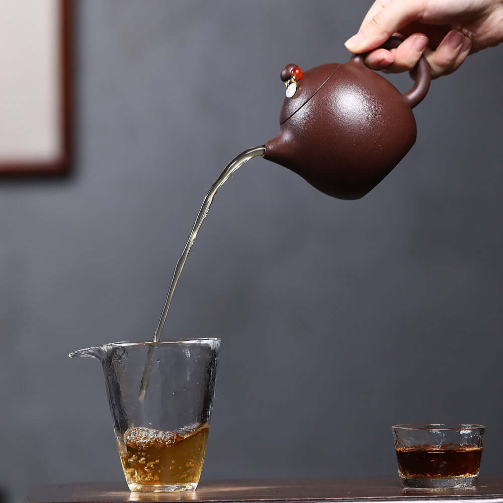 Handmade Yixing purple sand teapot pouring tea into a glass cup, showcasing unique craftsmanship in Kung Fu tea set.