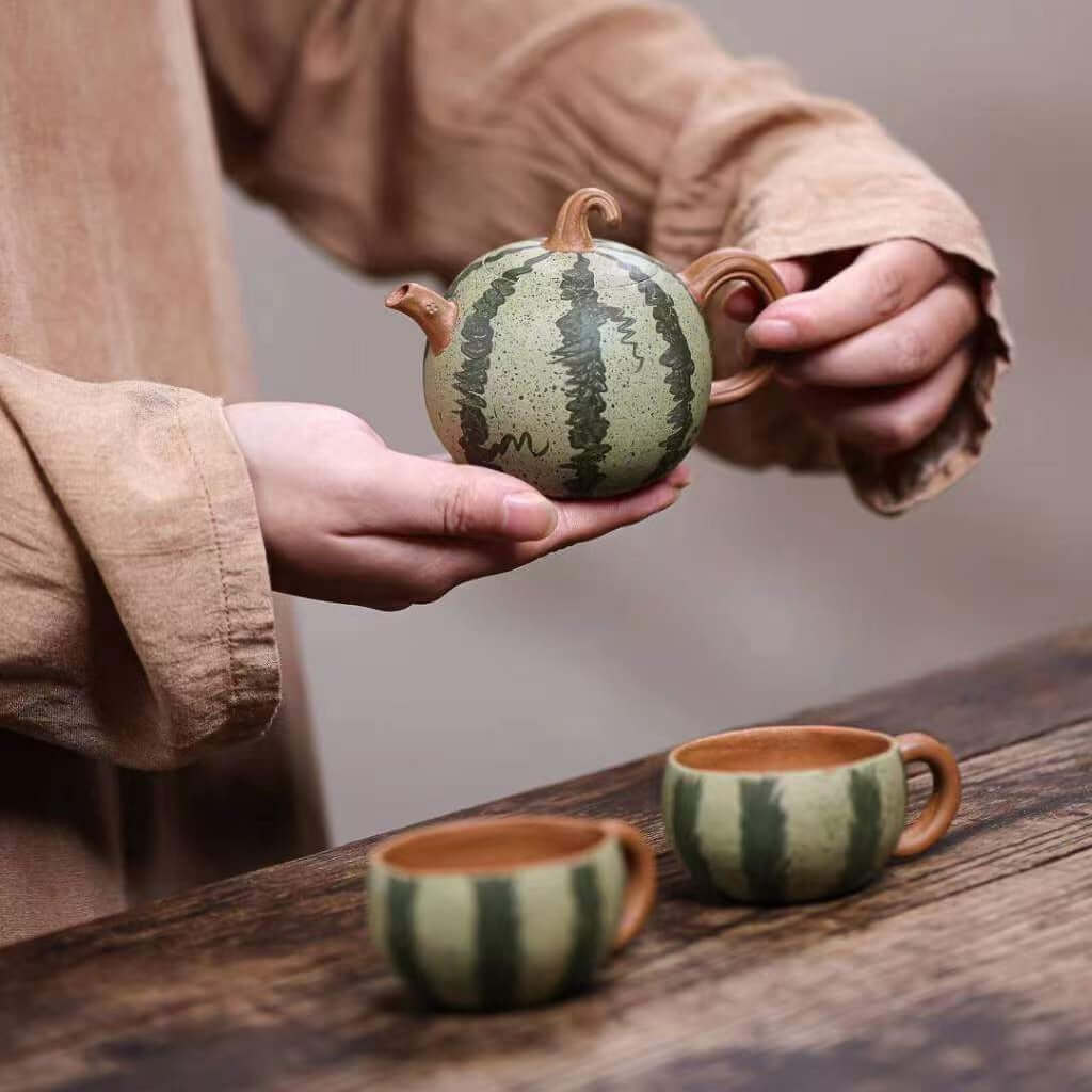 Handmade Yixing purple sand teapot in watermelon design, held in a hand; includes matching cups.