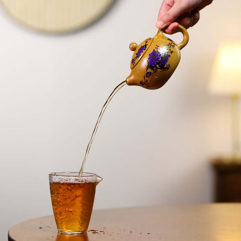 Handmade Yixing purple sand teapot pouring tea into a glass cup with warm amber liquid.