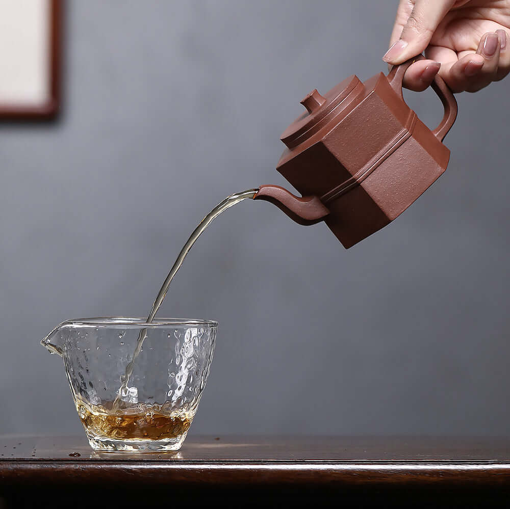 Handmade Yixing purple sand teapot pouring tea into a glass cup, showcasing its unique hexagonal design.