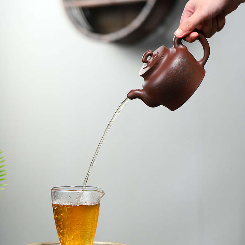 Handmade Yixing purple sand teapot pouring tea into a glass cup, showcasing elegant design and craftsmanship.