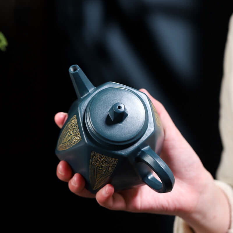 Handmade Yixing purple sand teapot, featuring intricate golden designs, held gracefully in a hand.
