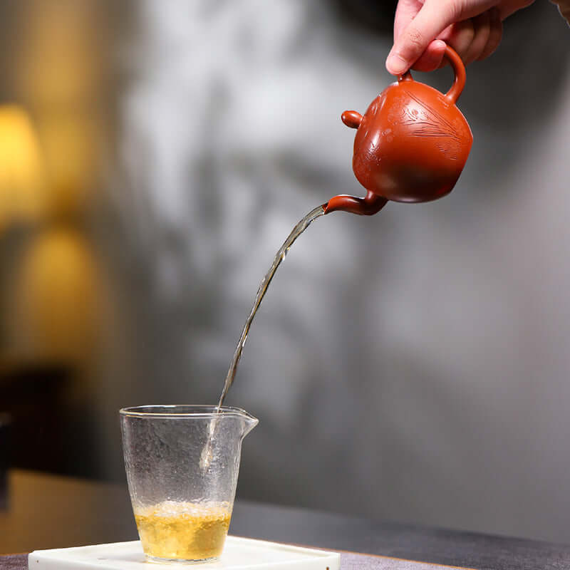 Hand pouring tea from a handmade Yixing purple sand teapot into a glass cup, showcasing traditional tea ceremony.