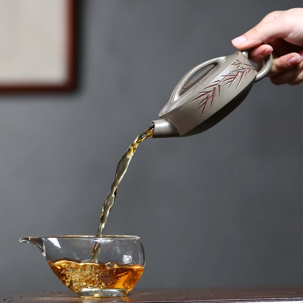 Handmade Yixing purple sand teapot pouring tea into a glass cup, showcasing traditional Kung Fu tea craftsmanship.