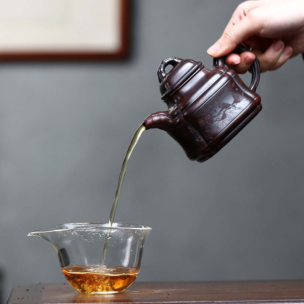 Handmade Yixing purple sand teapot pouring tea into a glass cup, showcasing exquisite craftsmanship and elegance.