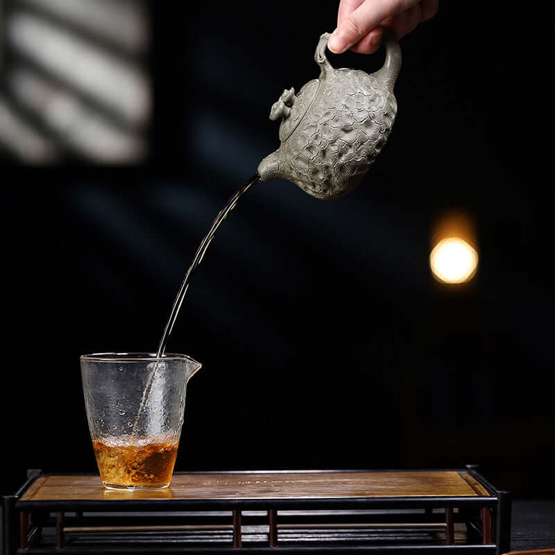 Handmade Yixing purple sand teapot pouring tea into a glass cup on a wooden tea tray, showcasing craftsmanship.