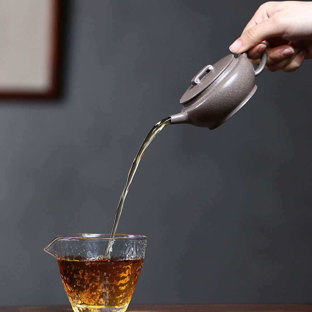 Handmade Yixing purple sand teapot pouring tea into a glass cup, showcasing traditional Chinese kung fu tea set craftsmanship.