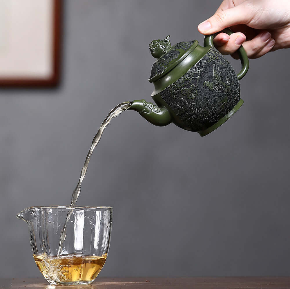 Handmade Yixing purple sand teapot pouring tea into a glass, featuring intricate flower and bird carvings.