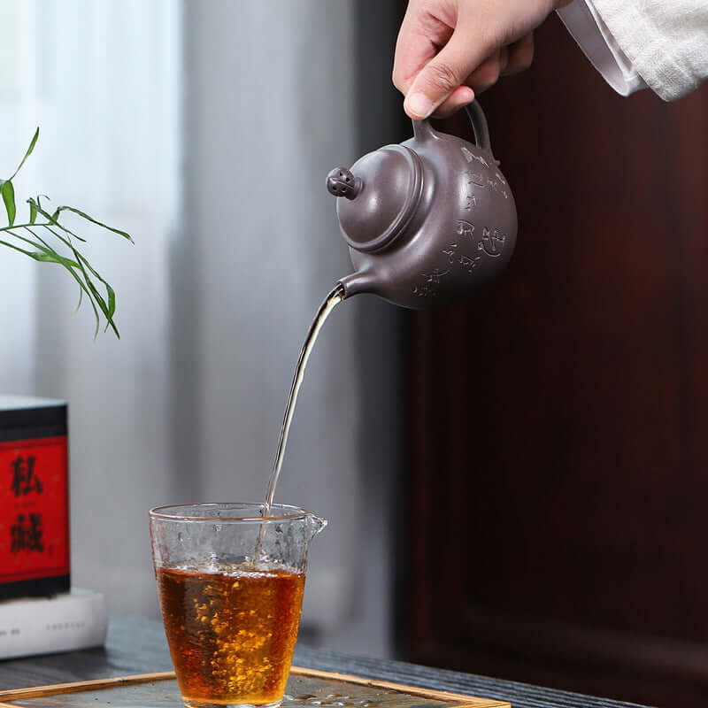 Handmade Yixing purple sand teapot pouring tea into a glass cup, showcasing traditional tea brewing elegance.